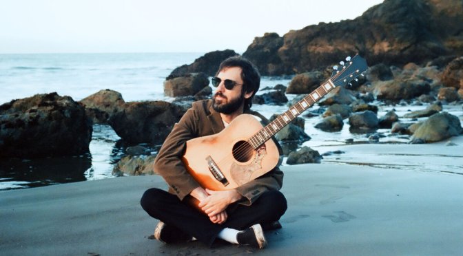 Rob Good of The Goods sitting on a beach holding an acoustic guitar