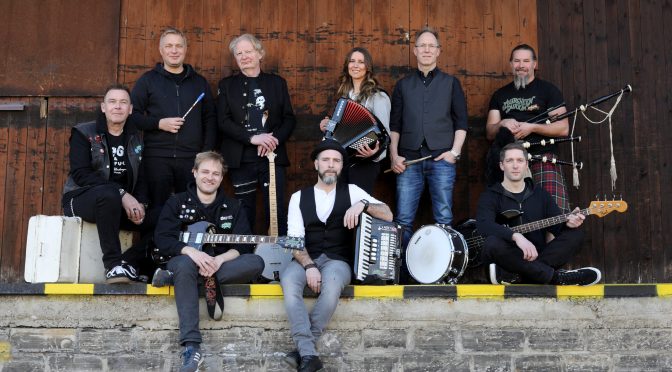 Muirsheen Durkin and Friends posing with bagpipes, accordion, banjo, guitars and drum in front of a wooden barn wall
